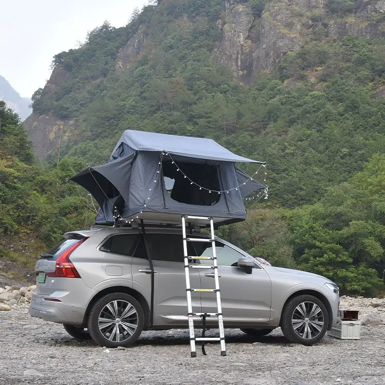 Silver SUV with grey soft shell rooftop tent opened, telescopic ladder, parked on gravel with mountain forest background.