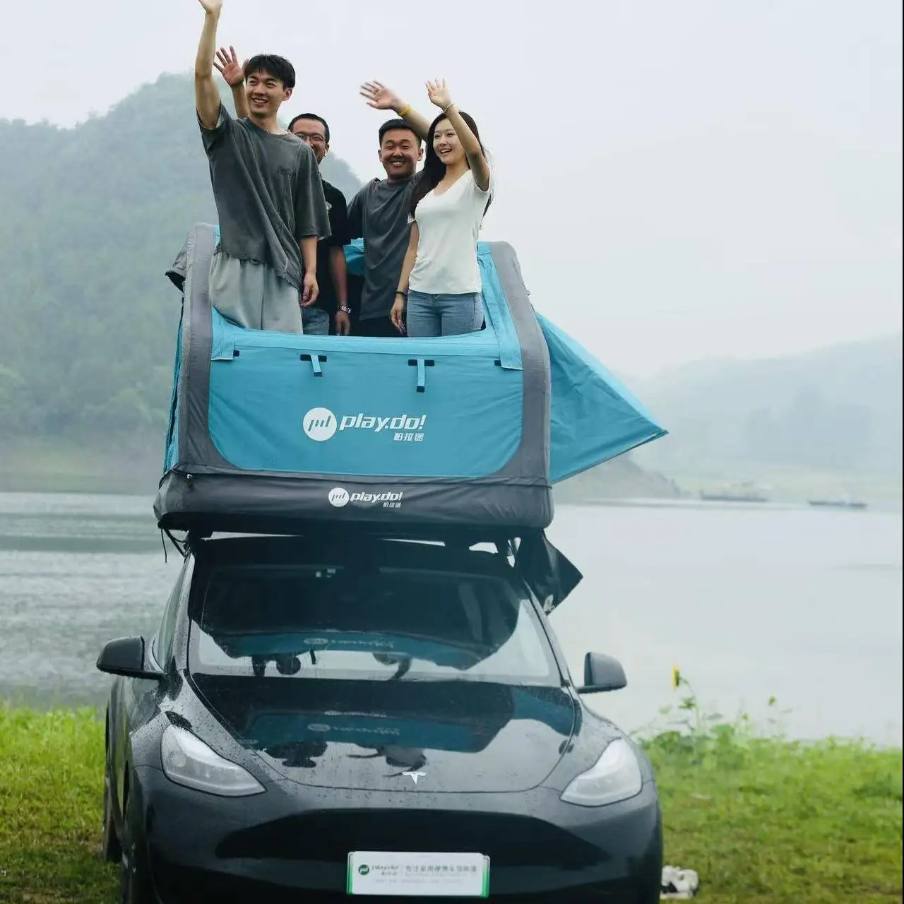 Group of campers waving from Fish Island rooftop tent on black SUV by lake, 2-3 person outdoor adventure